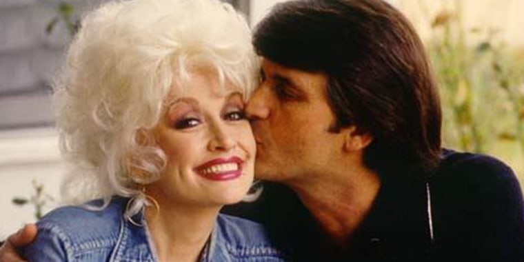 Dolly parton smiles as a dark haired main kisses her cheek.