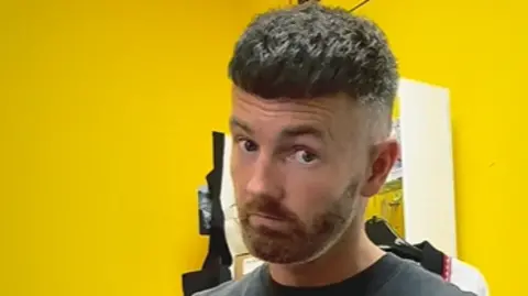Social media Chris has hair shaved close at the sides and longer on top. He is wearing a black vest and has his index finger raised towards the camera. Behind him is a yellow wall.