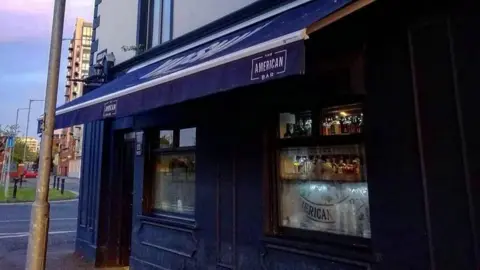 Social media The American bar is painted a dark colour, brown, almost black, and above it is a dark awning that hangs over the footpath.
