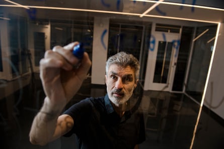 Yoshua Bengio holds up a hand to write in blue crayon on a glass surface. He has grey hair and a neat grey beard and wears a black top.