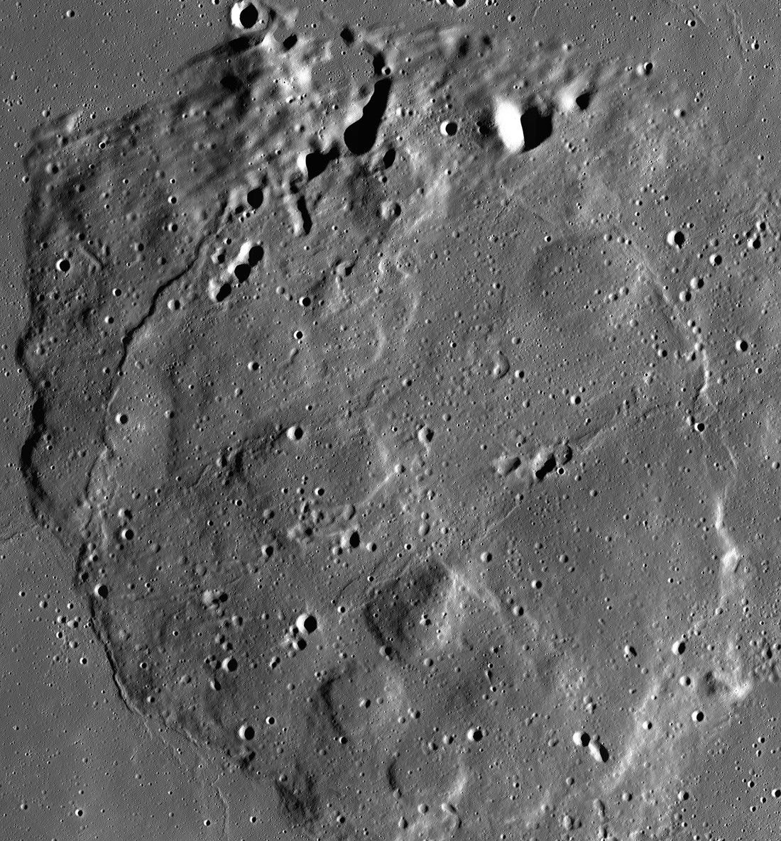 Overhead satellite mosaic showing Mons Rümker, a large, rounded volcanic mound on the Moon's surface surrounded by flat, dark lunar plains. The terrain is marked with impact craters of various sizes, with sharp sunlight casting deep, dark shadows along crater rims and the bumpy, elevated boundaries of the volcanic feature.